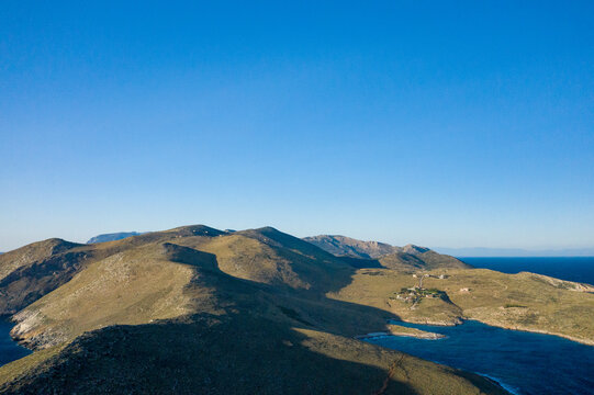 Aerial perspective of sunlit mountain ridges descending to a sheltered bay at Cap Tenare, Greece. Clear blue sky and rugged terrain evoke a sense of vastness and isolation.