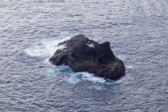 Isolated dark volcanic sea rock surrounded by vast ocean water and white foam waves.