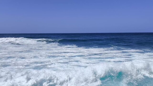 Slow motion ocean waves roll in with bright foam. Deep blue water swirls under a clear sky near the horizon.