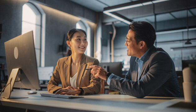 A professional man and woman collaborating at a modern office desk with a computer monitor and papers in a sleek, industrial-chic workspace.