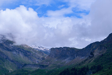 Fototapeta premium Steep rock faces rise above a lush green valley as low clouds drift overhead, with hints of snow and a waterfall visible in the dramatic mountain landscape.