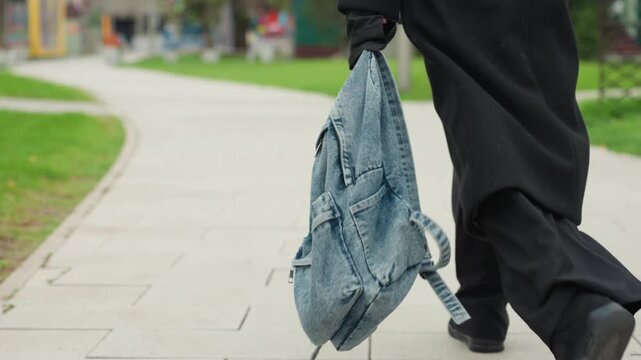 low angle unknown student walking with denim backpack, park pathway and green lawns, gentle casual stride, coat hem swaying, slow reflective mood, afternoon light, leisure campus scene