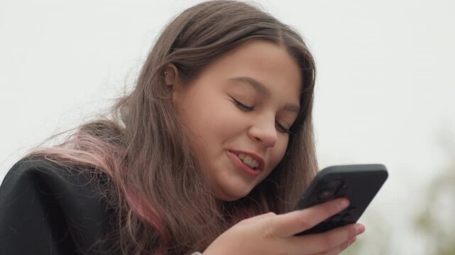latina student texting on smartphone outdoors, sending voice message and reading replies while smiling, pinkstreaked hair, wearing dark coat, overcast park background, candid closeup, relaxed