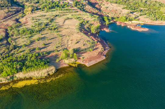 Aerial view of Lac de Salagou featuring rugged red shoreline, patches of green vegetation, and transparent blue water under bright sunlight.