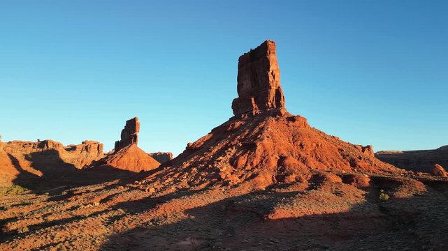 Drone shot in Valley of the Gods Utah USA showing Castle Butte and Stagecoach Rock fully illuminated by low early morning sun. Steady movement over red sandstone formations under clear blue sky.