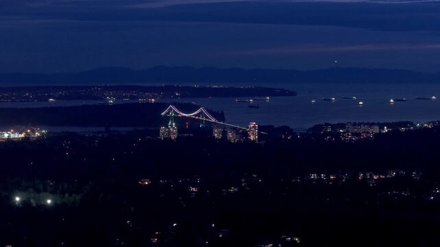 Elevated nighttime view of Vancouver Harbor featuring the illuminated Lions Gate Bridge, city lights, and anchored cargo ships across calm coastal waters under a deep blue sky.