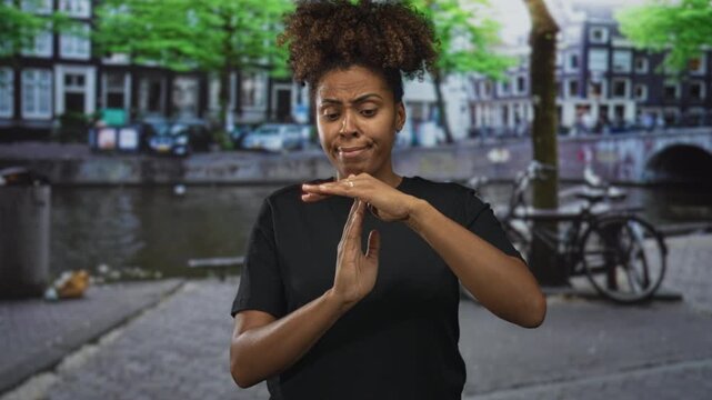 Woman with hands forming timeout sign and furrowed brow, wearing black t shirt near canal and bicycle on street in amsterdam; skepticism.