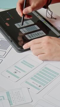 Vertical close-up of hands of anonymous UX designer choosing and ticking wireframes on graphic tablet screen, arranging smartphone application layouts on desk