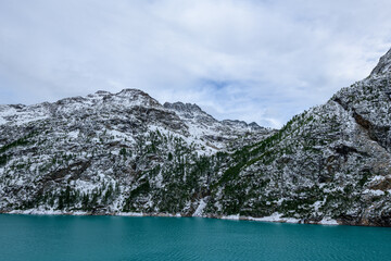 Obraz premium A vivid turquoise lake stretches beneath rugged, snow-dusted mountain slopes and scattered evergreen trees near Place-Moulin in the Aosta Valley, Italy. The dramatic alpine landscape is defined by