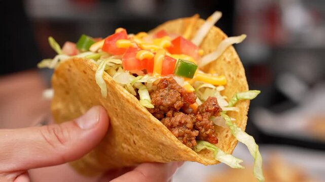 Hand Holding Crunchy Hard Shell Beef Taco Topped with Lettuce, Tomato, and Cheese Sauce | Delicious Mexican Fast Food Close-Up