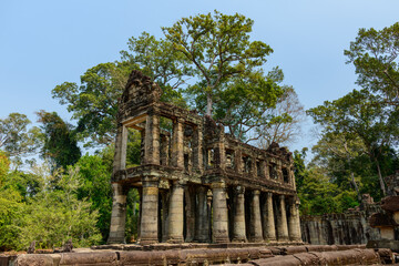 Obraz premium Two-story sandstone gallery with rows of columns stands amid dense tropical trees at Preah Khan temple in Cambodia's Angkor region. Weathered stone, mossy surfaces, and open blue sky evoke the