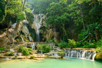Vivid green trees and dense tropical plants frame the terraced limestone cascades and turquoise pools of Kuang Si Waterfalls in northern Laos. Soft daylight highlights the flowing water and lush © Florent