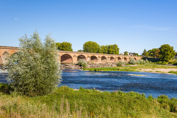 Fototapeta premium Historic stone bridge with multiple arches spans the Loire River under a clear blue sky in Nevers. The scene features flowing water, green riverbanks, and lush trees in bright summer light.