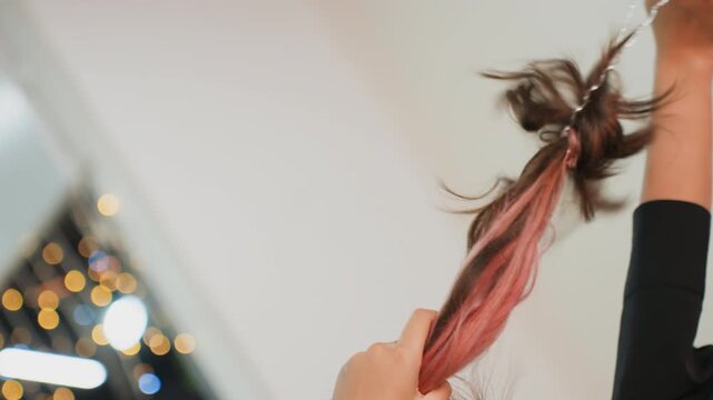 young student tying ponytail by bokeh lights, brunette with pink streaks adjusts hair in cozy indoor scene, hands styling through long tresses, warm soft glow and reflective mood, dorm window