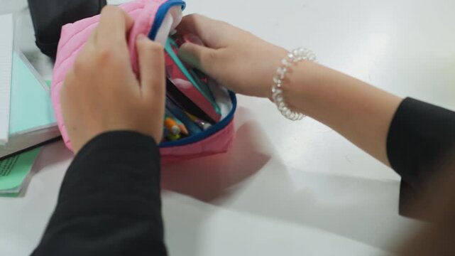 student hands opening pink pouch on desk retrieving compass and pencil focused study scene with tidy workspace, pastel accessories, bracelet on wrist, notebook and cup nearby, casual classroom