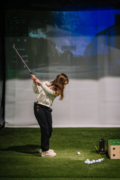 Young girl practicing golf swing indoors at a golf simulation area with artificial turf and digital screen in the background