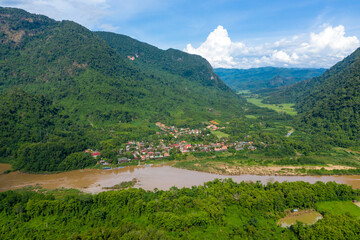 Fototapeta premium Aerial panorama of Muang Ngoi village nestled between steep green mountains and the muddy Nam Ou river in northern Laos. The scene features vibrant tropical vegetation, a broad river, and a dramatic