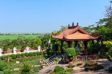 Obraz premium Red-roofed wooden pavilion surrounded by manicured shrubs and a lotus pond overlooks lush green countryside under a clear blue sky. The tranquil garden features sculpted statues and vibrant foliage in
