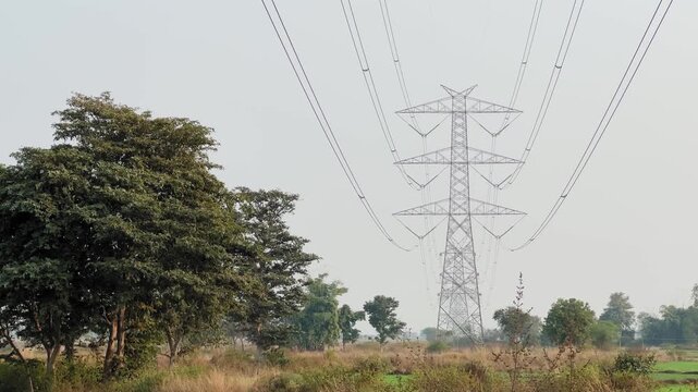 Tall 400kV transmission tower stands across rural fields, power lines curving overhead as trees and scrubland frame critical electricity infrastructure corridors network.