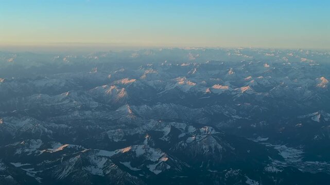 Elevated aerial view of snow-covered Alps mountains seen from a jet cockpit, with the scene bathed in sunrise light.