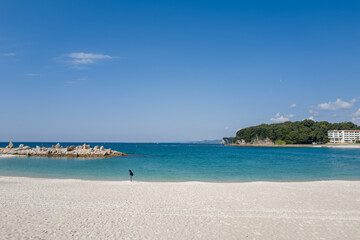A lone person stands on soft white sand facing turquoise water, with a rocky breakwater and lush green headland under a cloud-dotted blue sky in Shirarahama, Japan.