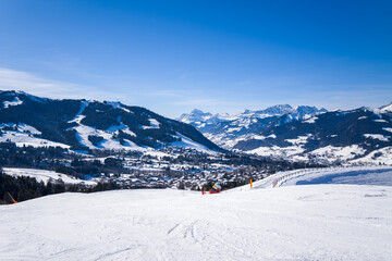 Wide view of snow-covered ski pistes overlooking Megeve village, surrounded by forested hills and distant alpine peaks under a clear blue sky. Bright sunlight enhances the crisp winter landscape and