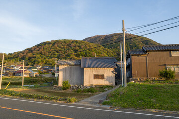 Obraz premium Wooden rural homes stand along a quiet roadside, backed by lush green hills under warm evening sunlight. The scene captures a peaceful village atmosphere with natural textures and soft shadows.