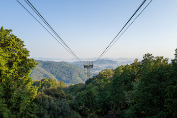 Suspended cable car lines extend above a lush green valley with dense trees, leading toward distant cityscape and hazy mountains near Himeji, Japan. Soft daylight creates a tranquil, expansive