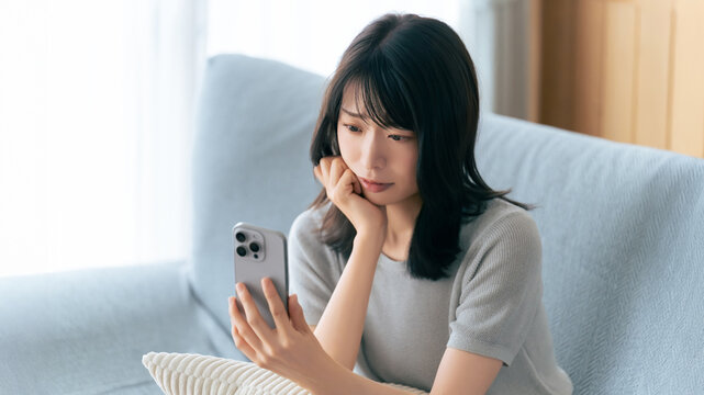 スマートフォンを見ながら考え込む女性,自宅のソファで過ごす時間, A Woman Looking Thoughtful While Checking Her Smartphone on a Sofa at Home