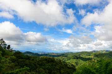 A wide view of lush green hills and dense forest stretches beneath a sky filled with large white clouds and patches of vibrant blue. The landscape captures the natural beauty and rolling terrain of