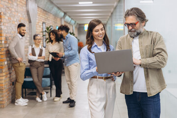 Business people collaborating on a laptop in a modern office hallway, discussing work, teamwork and mentoring
