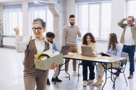 Mature woman happily exiting her corporate job, holding a box of personal items, colleagues showing various reactions