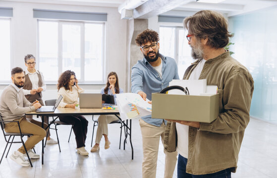 Employee leaving workspace, carrying personal belongings in a box, colleague handing over final documents, team watching goodbye