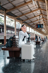 Smiling woman with headphones and suitcase at a train station, ready for a journey.