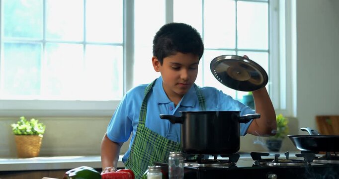 Indian asian young kid cooking tasty soup in pot and making father taste with wooden spoon while smiling together in modern kitchen, playful family bonding and learning moments at home