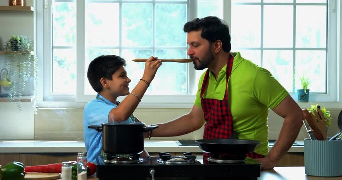 Indian asian young kid cooking tasty soup in pot and making father taste with wooden spoon while smiling together in modern kitchen, playful family bonding and learning moments at home