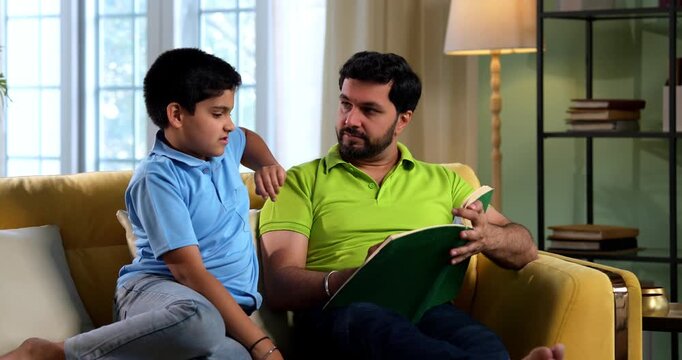 Indian asian father and son reading a book on sofa in modern home while dad explains school chapter and tells story to small kid sitting together in cozy living room family education bonding moment