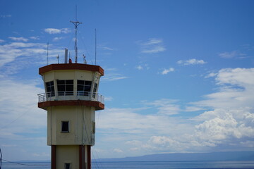 A modern airport control tower standing tall against a clear blue sky with soft white clouds and a distant ocean horizon. 