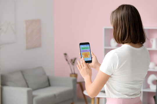 Beautiful young woman using mobile phone with smart home security system control panel in room, back view