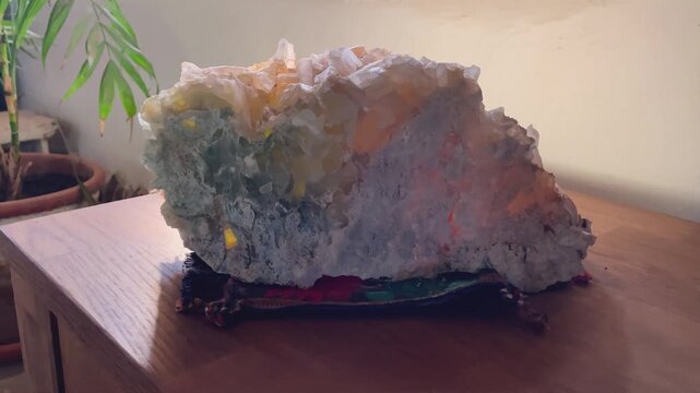 Slow motion footage of a large lapis specularis crystal stone with greenish and orange tones. The mineral rests on a decorative fabric over a wooden surface, partially illuminated by soft window light