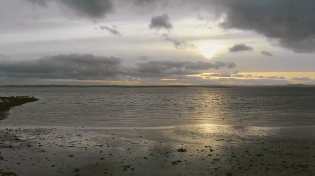 Aerial dolly left over a Belfast calm coastal shoreline with shallow water, tidal flats, distant landforms, and an overcast sky.