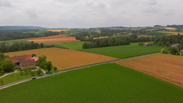 Aerial wide shot of rural farmland with patchwork fields, country road, scattered houses, and rolling hills under an overcast sky.