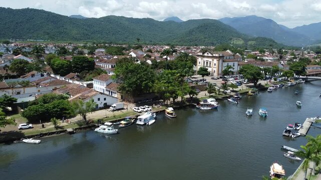 Drone shot crossing Rio Pereque Acu in Paraty, Brazil, capturing tourist boats, and jet skis on the river. Features the town, Igreja Nossa Senhora dos Remedios, mountains, and Atlantic forest