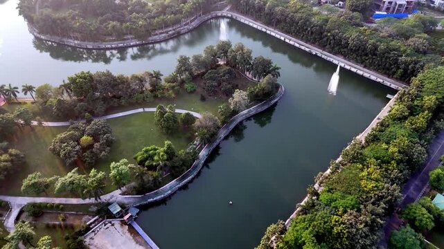 Drone shot of a serene city lake featuring winding pathways, tree-covered islands, and water fountains, showcasing planned urban green spaces and waterfront.
