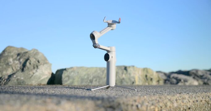 Smartphone gimbal stabilizer set up as a tripod on rocks against a clear blue sky. Ground level pov