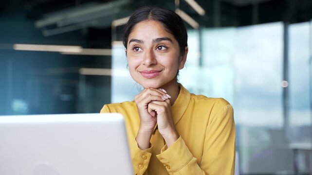 Happy dreamy young businesswoman sitting at desk in modern office resting her chin on hands. Smiling female employee looking away with positive thoughts, enjoying moment of inspiration at workplace