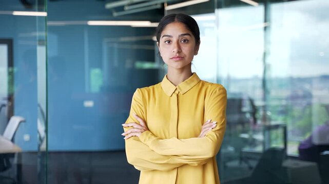 Portrait of young serious businesswoman standing with crossed arms in modern glass office. Confident professional female in yellow blouse looking at camera, posing at workplace with determination.
