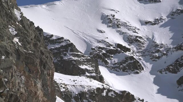 Panoramic snowy mountain range shows jagged rocky peaks, ridgeline contours, clear sky. Wide cinema vista reveals ice covered highland summits, crags, crest forms, azure backdrop.