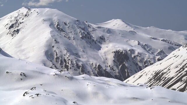 Panoramic snowy mountain range shows jagged rocky peaks, ridgeline contours, clear sky. Wide cinema vista reveals ice covered highland summits, crags, crest forms, azure backdrop.