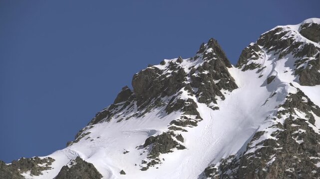 Panoramic snowy mountain range shows jagged rocky peaks, ridgeline contours, clear sky. Wide cinema vista reveals ice covered highland summits, crags, crest forms, azure backdrop.
